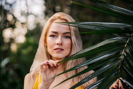 Beautiful girl with natural makeup, blonde hair standing in rainforest among exotic trees. Closeup portrait of magnificent woman in yellow swimsuit among tropical plants. Focus on beautiful palm leavesの写真素材