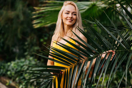 Young sexy woman with slim body, wearing yellow swimsuit standing among tropical trees. Horizontal shot of charming girl smiling posing in green leaves during holiday at resort. Summer, vacation.の写真素材