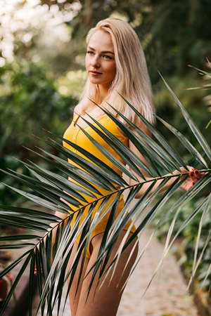 Close up vertical portrait of happy beautiful luxurious blonde with cute smile standing near palm tree on the beach, wearing bright swimsuit, relaxing on tropical island. Vacation, travel, summer.の写真素材