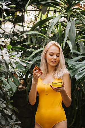 Beautiful caucasian woman with healthy smooth clean skin holding jar of body cream, sunblock, smelling its scent with closed eyes, posing in rainforest. Skincare, spa, natural cosmetic product conceptの写真素材