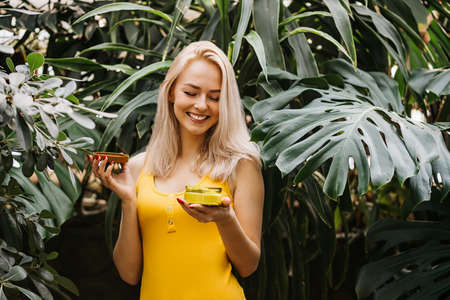 Beautiful caucasian blonde woman with healthy smooth clean skin holding jar of body cream, applying sunscreen, standing on tropical green leaves background. Concept of natural cosmetics and skin care.の写真素材