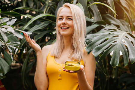 Beautiful caucasian blonde woman with healthy smooth clean skin holding jar of body cream and a happy contented face sniffs it against the background of exotic woods. cosmetic product conceptの写真素材