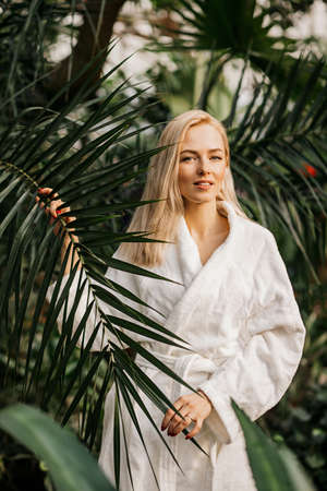 Happy lovely cheerful female model wearing white bathrobe standing behind palm leaf, looking to side and smiling. Spa, treatment, natural cosmetics, skin care, cosmetology concept.の写真素材