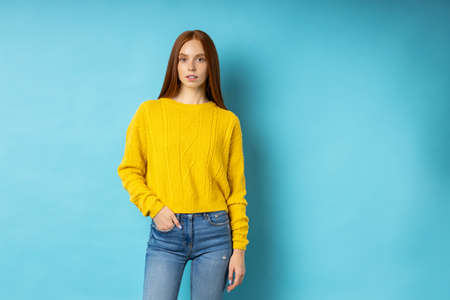 Portrait of redhead attractive slim caucasian serious calm young lady wearing yellow sweater, jeans keeping hand in pocket posing on blue background with copy space.の写真素材