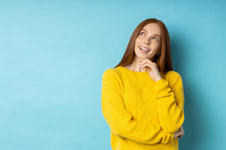Dreamy cute redhead young woman looking thoughtfully aside, touching chin with hand dreaming about vacation, planning something isolated on light blue background with copy space.の写真素材