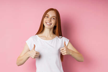 Portrait of redhead female student with broad smile, looking at camera with happy expression, showing thumbs up with both hands, showing her approval standing against pink studio wall. Body languageの写真素材