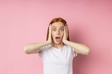 Terrified amazed caucasian woman looking with bugged eyes, open mouth, keeping palms on head, being...