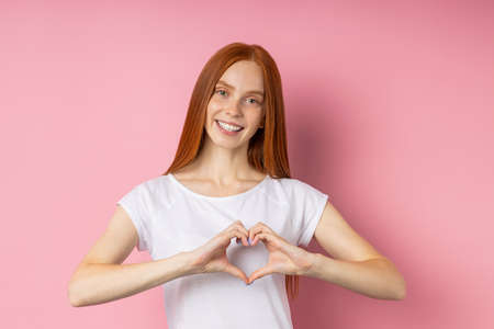 Portrait of beautiful red haired woman with freckled face, showing heart gesture, confessing in love, having romantic feelings, wearig white t shirt smiling broadly standing over pink wall.の写真素材