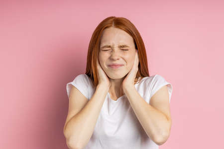Closeup portrait of stressful young redhead freckled woman covering ears with palms and closing eyes tight,annoyed with loud noise, having headache, migraine. Expressive facial expressions. Copy spaceの写真素材