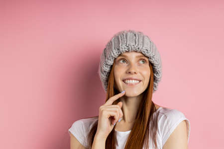 Beautiful young ginger woman with no makeup keeping hands under chin, wearing winter knitted beanie, looking aside with dreamy happy expressions isolated on pink wall. People, good emotions concept.の写真素材