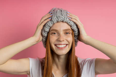 Excited surprised redhead woman with freckled clean skin in white t shirt, trendy beanie, with opened mouth, touching hat with both hands, reacting on something awesome isolated over pink backgroundの写真素材