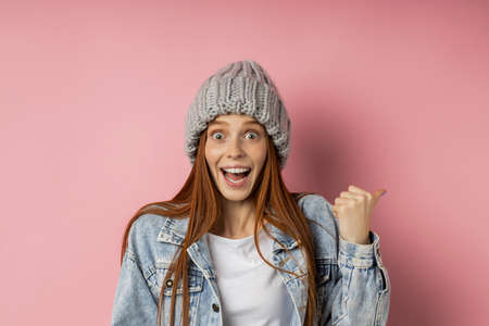Amazed joyful female model with red hair, wearing gray knitted bulk hat, looking at camera with bugged eyes and opened mouth, pointing with finger to side posing on pink background with copy space.の写真素材