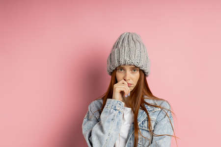 Confused puzzled carefree young caucasian ginger woman in gray knitted hat, denim jacket shrugging shoulders looking at camera with guilty and uncertain facial expression isolated pink background.の写真素材