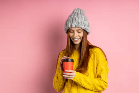 Shot of happy caucasian woman with long red hair holding takeaway coffee, enjoying break, smiling with white teeth looking at camera posing against pink background with copy space for advertising.の写真素材
