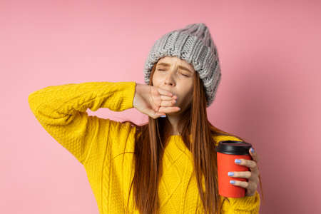 Overworked caucasian redhead woman dressed in yellow sweater, knitted gray hat, having sleepy and tired look, yawning covering mouth with palm, holding takeaway coffee in hand, posing over pink wall.の写真素材