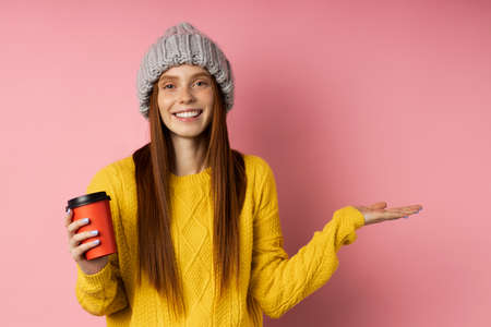 Optimistic happy girl wearing casual clothes holding copy space in one hand, disposable cup of hot drink in other hand, keeping palm open smiling posing on pink background.の写真素材