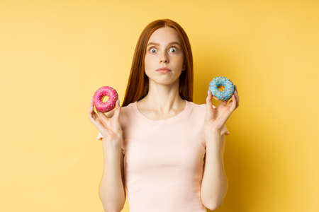Unhappy frustrated slim ginger young woman on diet holding two color donuts, cant eat delicious confectionery, isolated over yellow background. Junk food, dieting, weight loss concept.の写真素材