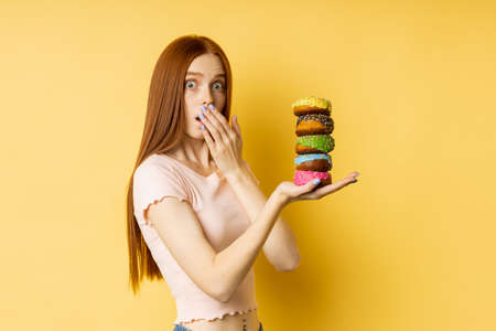 Shot of slim nice surprised caucasian redhead woman in casual t shirt holding pile of colorful donuts, looking with puzzled expression over yellow background. Bakery, unhealthy food, diet concept.の写真素材