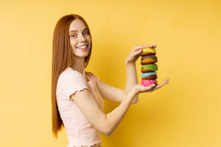 Happy cheerful redhead girl sweet tooth holding pile of colorful glazed donuts, broadly smiling looking at camera against yellow studio wall. Junk food, pastry, bakery concept.の写真素材