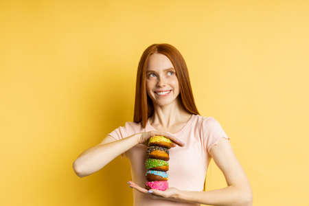 Happy joyful redhead woman sweet tooth holding pile of colorful glazed donuts, broadly smiling looking at camera standing against yellow studio wall. Pastry, bakery concept.の写真素材