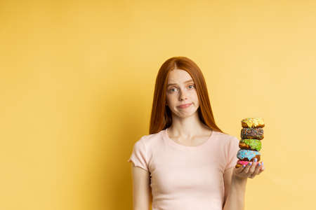 Unhappy frustrated slim ginger young woman on diet holding color delicious donuts, cant eat pastry, isolated over yellow background with copy space. Junk food, dieting, weight loss concept.の写真素材