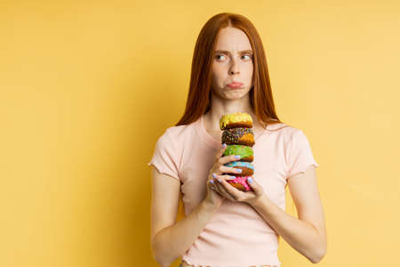Waist up shot of serious caucasian woman with long red hair, in t shirt holding colorful glazed donuts, looking strictly at camera over yellow background. Unhealthy food, dieting, concept.の写真素材