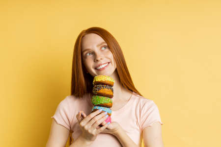 Horizontal closeup shot of pleased redhead woman holding colorful decorated donuts, broadly smiling looking at camera, enjoying sweets, isolated yellow background with copy space for your advertising.の写真素材