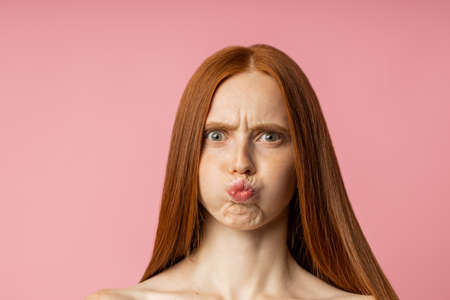Attractive caucasian red long haired naked woman frowning eyebrows, making faces, pursing lips looking at camera standing over pink background. Facial expressions, emotions concept.の写真素材