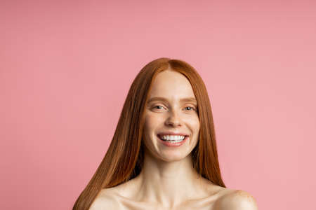 model with shiny straight long red hair. Cheerful girl with clean fresh skin pleased with hair treatment, smiling at camera posing on pink background. Keratin straightening. Spa procedures.の写真素材