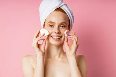 Pretty glad young caucasian woman with towel on head, bare shoulders, holding two cosmetic sponges on cheeks, enjoying beauty day smiling broadly posing against pink studio wall. Skin care.の写真素材