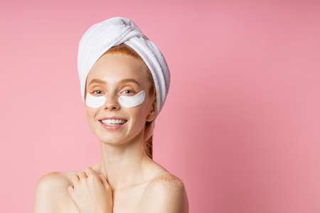 Closeup of beautiful happy caucasian woman with moisturizing under eye mask, towel on head, fresh clean skin, bare shoulders, cute smiling at camera isolated on pink background. Under eye patches.の写真素材