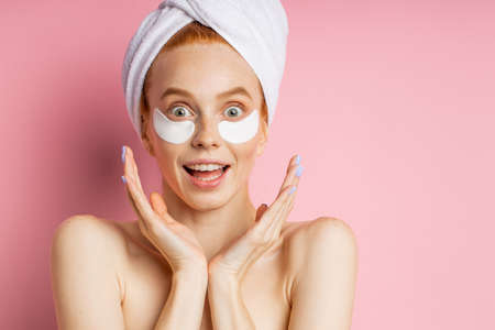 Closeup of surprised cheerful redhead young woman applying moisturizing hyaluronic patches, biting lips, looking to side with happy expression posing over pink background with copy space.の写真素材