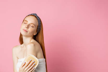 Skin care, hygiene concept. Cropped shot of attractive sensual caucasian girl using washcloth, posing with closed eyes, wrapped in white towel, enjoying body care treatment, isolated over pink wall.の写真素材