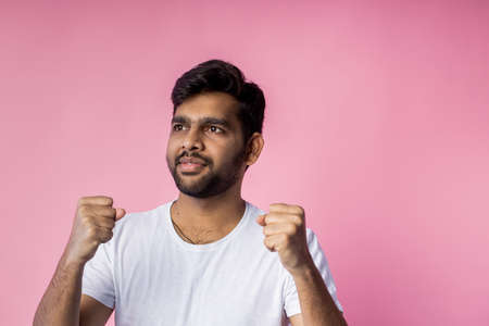 People, success, win, good news and celebration. Happy successful young indian male raising hands, clenching fists in triumph, celebrating victory, wearing casual white t shirt, on pink background.の写真素材