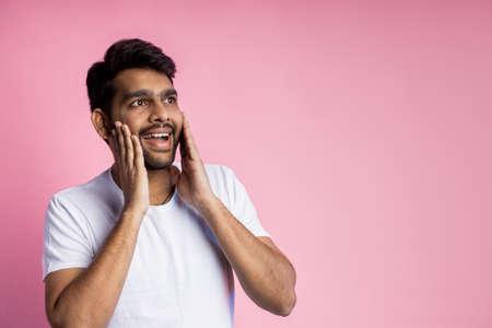Portrait of pleasantly surprised happy man in white t shirt, touching cheeks with palms, looking aside with amazed expression, isolated on pink background with copy space for advertising or text.の写真素材