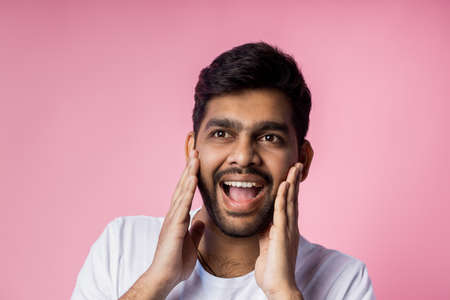 Stunned handsome unshaven indian male, being shocked by low prices on sale, keeping hands on cheeks, opening mouth in surprise, isolated over pink background. Emotions, reactions concept. Copy space.の写真素材