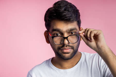 Handsome smart indian guy, student dressed in white t shirt, smiling looking at camera, wearing glasses and touching it, posing in studio over pink background. Facial expressions concept.の写真素材