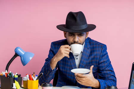 office worker, businessman in formal checkered suit, black hat, sitting at workplace, thinking about something, looking aside, enjoying aromatic coffee. Business, lifestyle, people concept.の写真素材