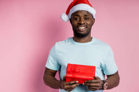 Handsome happy african american male wearing Santa hat, holding red gift box, smiling, giving present to girlfriend, isolated over pink background. Christmas concept.の写真素材