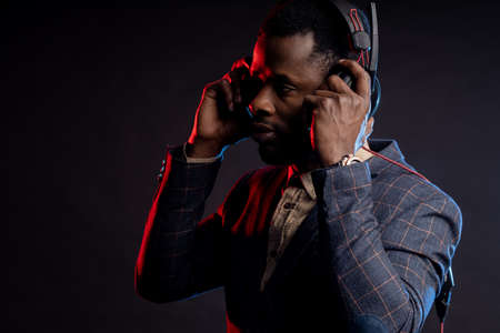 Black man with headphones on the side, listening music, posing in dark. Portrait of stylish DJ with headset, wearing checked suit, isolated on black backgroundの写真素材