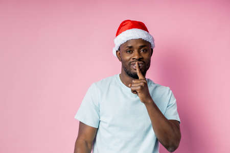 Confident young  male with Santa hat on head, making shh gesture, keeping index finger on lips, telling secret, asking for silence, isolated over pink background.の写真素材