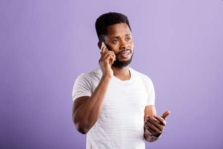 Serious sad african american man talking on mobile phone, wearing white tshirt looking up with thoughtful dissatisfied expression on violet background. Face expressions and communication concept.の写真素材