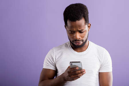 Photo of amazed puzzled black man holding modern cell phone, receiving unexpected message, reading shocking news, isolated over lilac background. Reaction, technology, facial expressions concept.の写真素材