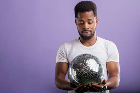 Happy dark skinned man holding shining disco ball, going to party with friends, celebrating...