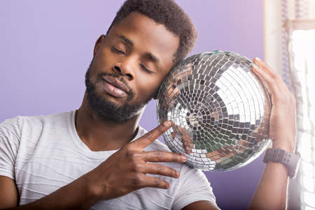 Handsome relaxed unshaven african american man holding shining disco ball, enjoying with closed eyes dreaming about party wearing white tshirt posing over violet background. Close up.の写真素材
