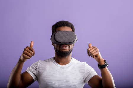 Dark skinned young man experiencing virtual reality, showing thumb up, playing video games using oculus rift headset. 3d technology, innovation, entertainment, cyberspace concept.の写真素材