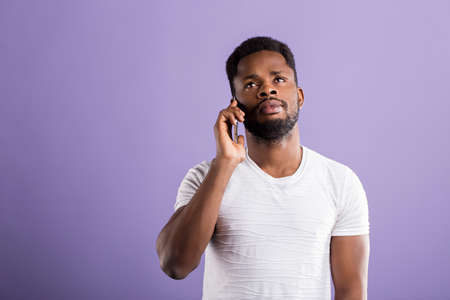 Serious sad african american man talking on mobile phone, wearing white tshirt looking up with thoughtful dissatisfied expression on violet background. Face expressions and communication concept.の写真素材