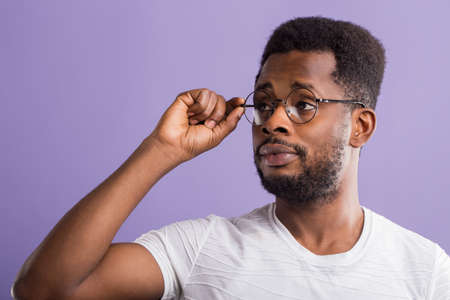 Close up of surprised young handsome african american guy with short curly hair and small beard touching spectacles looking attentively to side, has bad eyesight isolated on purple background.の写真素材