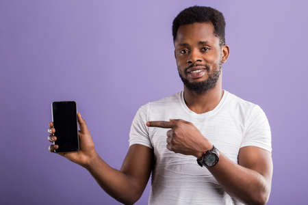 Positive happy african guy dressed in casual outfit, showing modern black smartphone and pointing at device with index finger, smiling over violet studio wall. Technology, communication concept.の写真素材