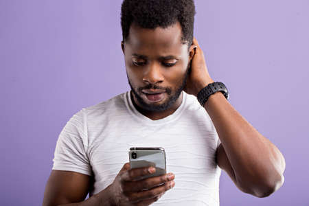 Photo of amazed puzzled black man holding modern cell phone, receiving unexpected message, reading shocking news, isolated over lilac background. Reaction, technology, facial expressions concept.の写真素材